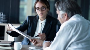 Une femme au look professionnel, vêtue d'un costume, montre un document important à un homme portant une chemise blanche. Le visage de la femme est visible, tandis que celui de l'homme est obscurci.