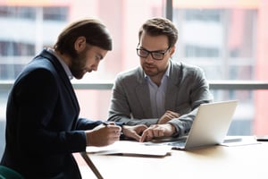 Deux hommes sont assis côte à côte au coin d'une table, examinant des documents importants avec un ordinateur portable ouvert. Un homme signe avec un stylo tandis que l'autre montre où signer.