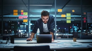 A man is standing and leaning on a table, looking at his laptop in a conference room. Colorful post-its on the glass walls suggest a brainstorming session took place, but now the man is alone. The city behind him is dark, indicating he is working late into the night..