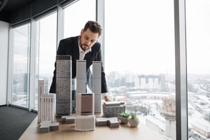 Un homme regarde une maquette d'un ensemble de gratte-ciels et d'autres bâtiments. Il a une vue sur une ville à travers les fenêtres du sol au plafond de son bureau.