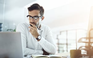 A man is sitting at a desk, looking intently at a computer screen. He is wearing glasses and has a stylish watch on his wrist.