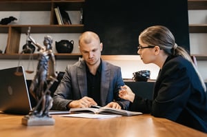 Un homme et une femme regardent un livre à une table, qui semble être un manuel de droit. Devant eux, sur la table, se trouve une statue de la Justice placée sur le côté.