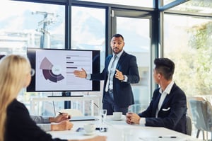 A man is presenting in a boardroom, standing in front of a chart that displays statistics and figures about their company. At least three other people are present, with two visible and one obscured. 