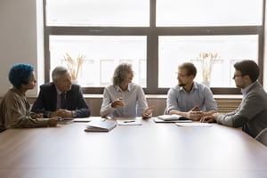 Un groupe de professionnels est assis d'un côté d'une table, vu de face. Le groupe présente une diversité, avec des individus comme un professionnel plus alternatif aux cheveux bleus, aux côtés de professionnels plus âgés vêtus de costumes, mettant en avant un mélange de styles et d'expériences.