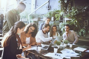 Un groupe de professionnels est assis autour d'un bureau rempli d'ordinateurs et de papiers, se concentrant sur leur travail et collaborant ensemble. L'arrière-plan est orné de nombreuses plantes, créant une ambiance de bureau axée sur l'humain et accueillante.